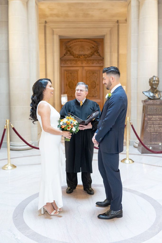 Couple getting married in a civil ceremony on the Rotunda inside SF City Hall, smiling at each other with smiling officiant