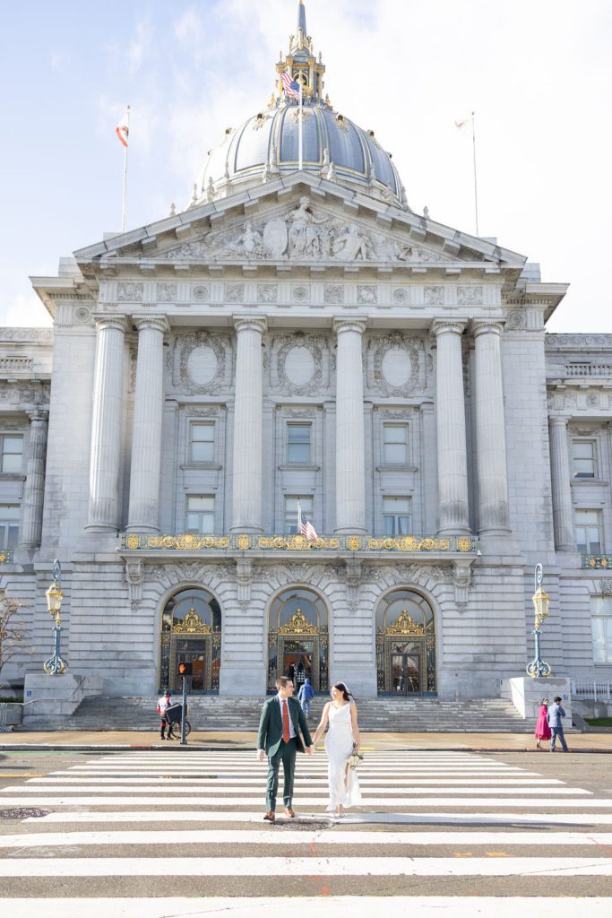 Front of San Francisco City Hall on a mostly sunny day, with newly married couple walking across crosswalk holding hands and smiling at each other