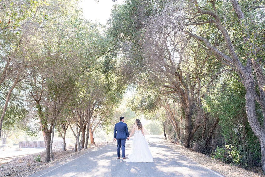 Nicole and Daniel, holding hands and walking on a tree-lined path at Sunol's Casa Bella during their wedding.