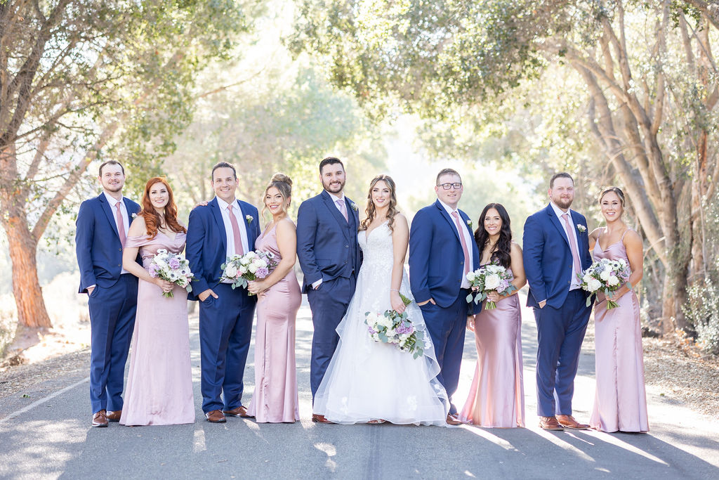 A wedding party, Nicole and Daniel's Wedding, posing on a tree-lined pathway in Sunol's Casa Bella, with men in suits and women in formal dresses holding bouquets.